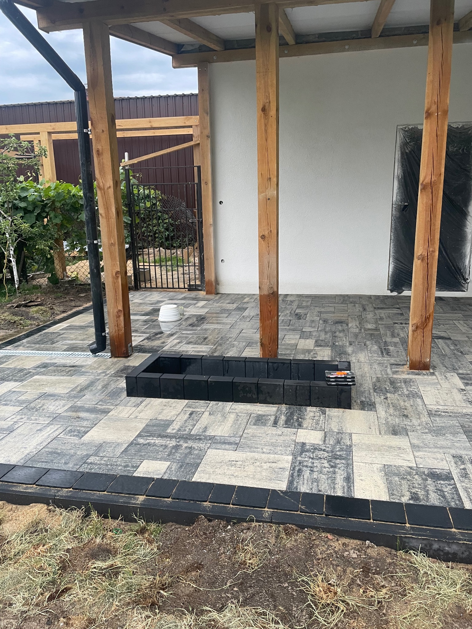 Wykończony taras z kostki brukowej w odcieniach szarości i czerni, z czarną obwódką i elementem dekoracyjnym w kształcie kwadratu. Widoczne drewniane podpory zadaszenia.