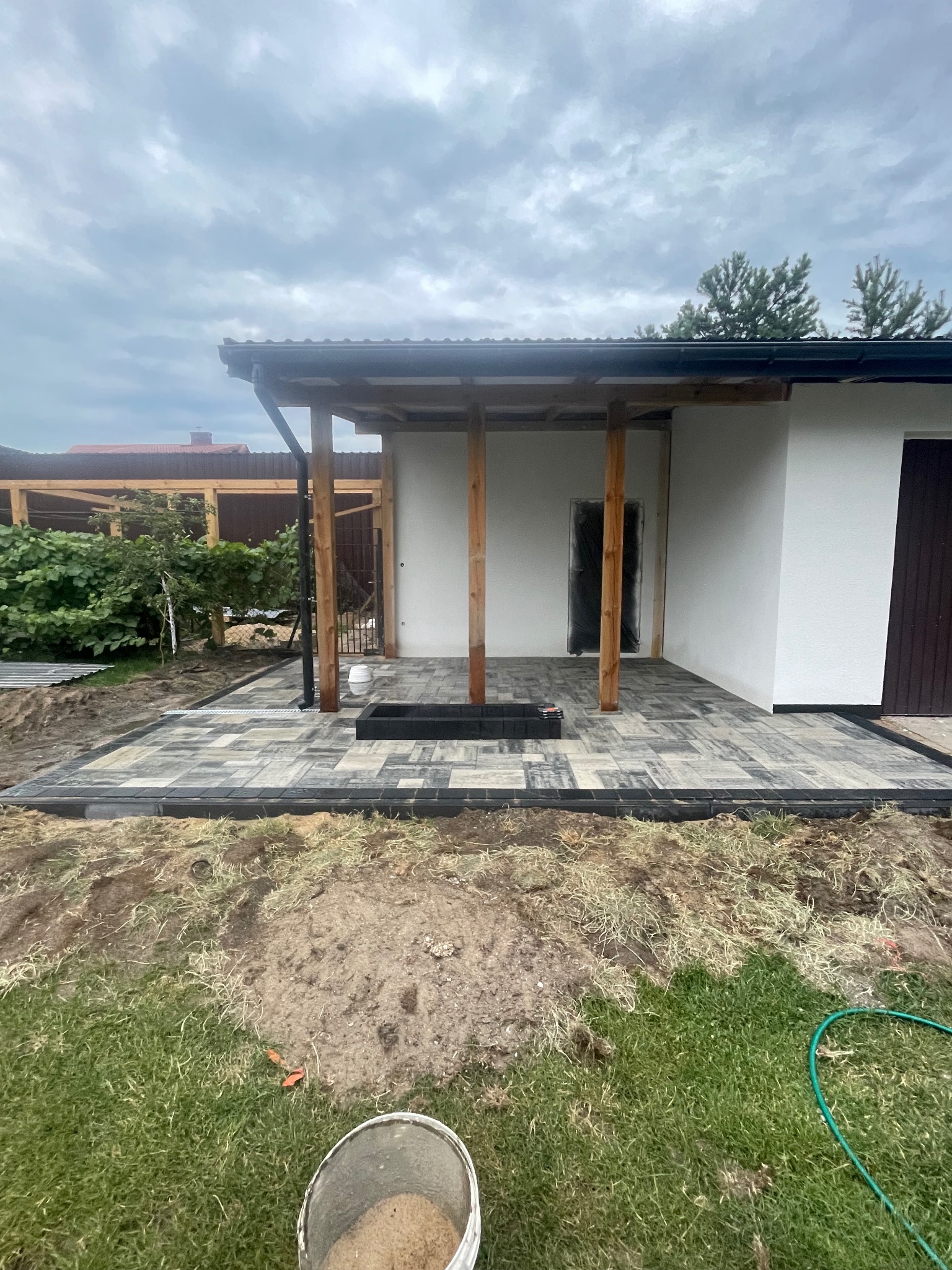 Ułożona kostka brukowa na tarasie z drewnianą konstrukcją zadaszenia, widok na ogród i dom. Kostka w odcieniach szarości, czarna rynna, wiadro z piaskiem w rogu kadru.