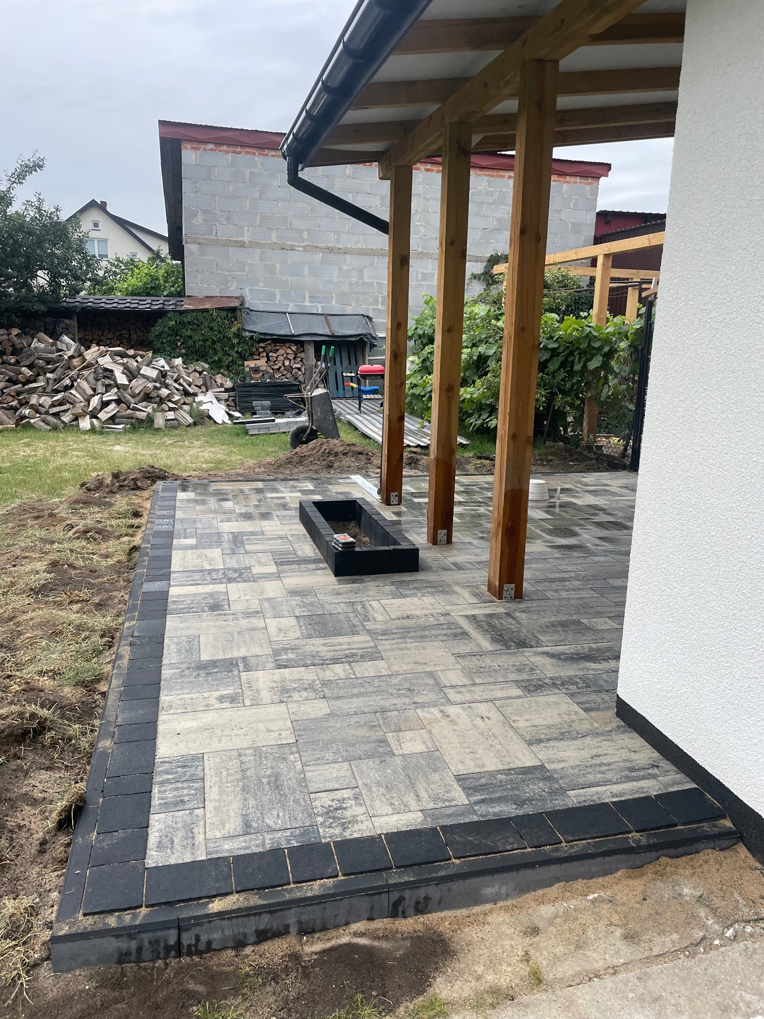 Nowoczesny taras z kostki brukowej w odcieniach szarości, obramowany czarną kostką. Widoczne drewniane słupy zadaszenia i palenisko. W tle budynek i sterta drewna.