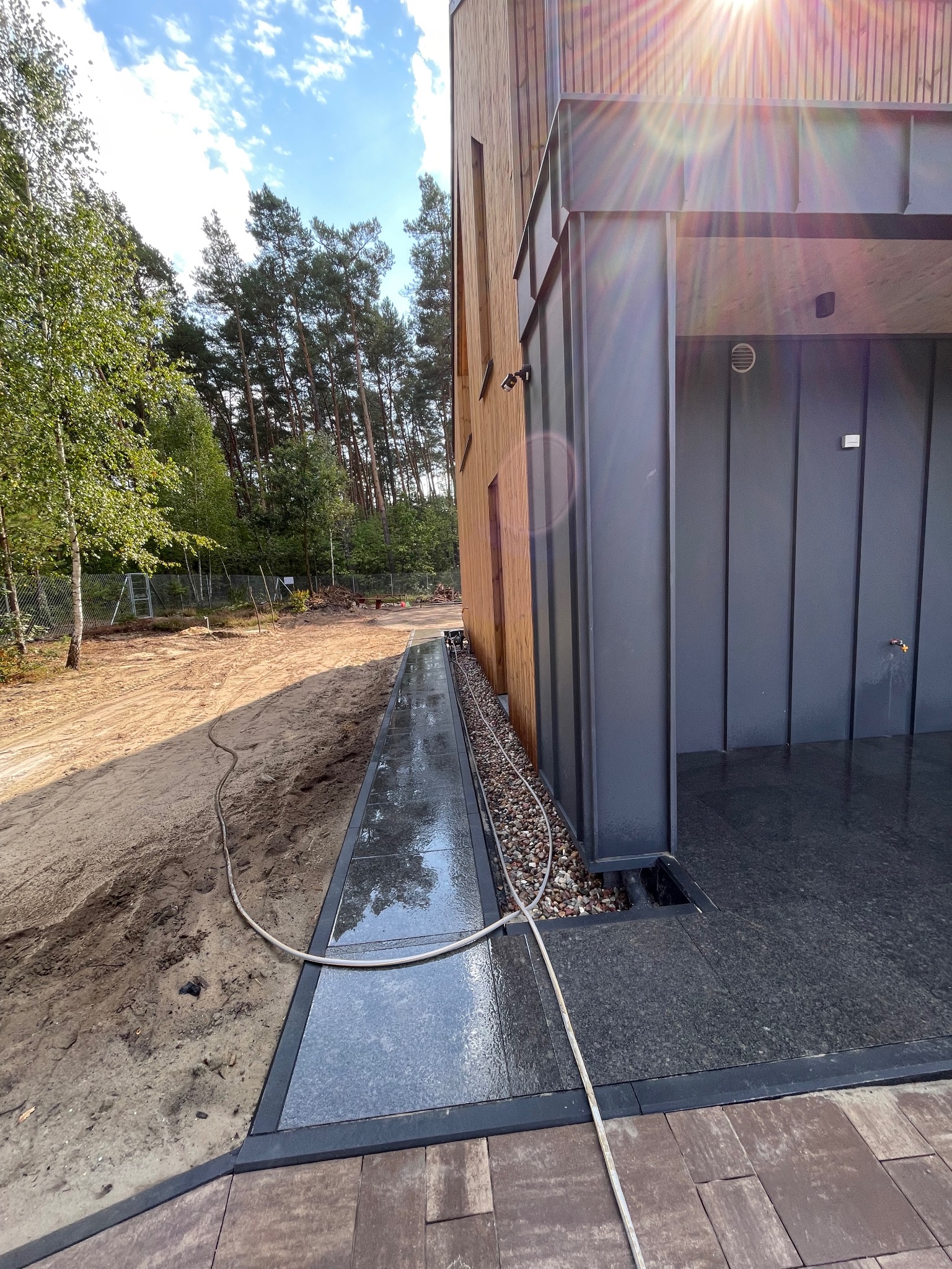 Nowoczesny drenaż liniowy z kamienia naturalnego przy budynku z drewnianą elewacją. Widoczny wąż ogrodowy i teren przygotowany pod dalsze prace.