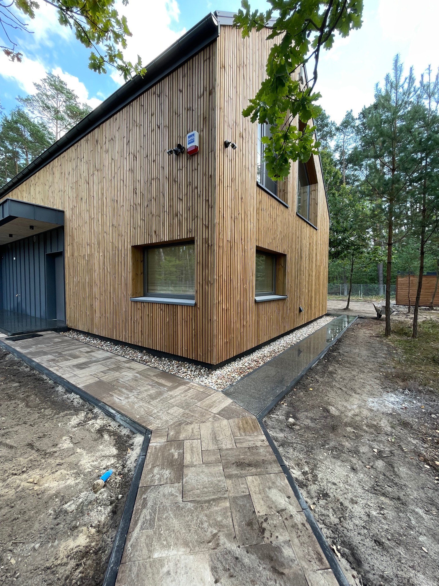 Nowoczesny dom z drewnianą elewacją i ścieżką z kostki brukowej. Ujęcie z poziomu gruntu, widoczne detale wykończenia i otoczenie domu.