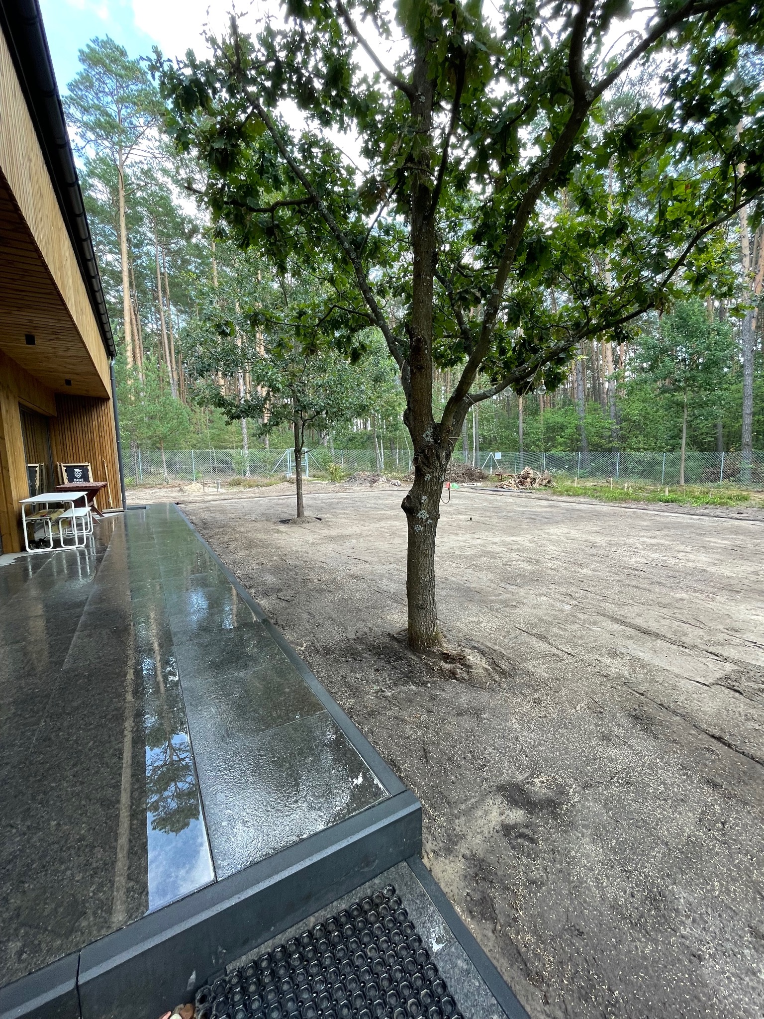 Przygotowany teren pod budowę domu z wykończonym tarasem z ciemnej kostki, drzewa na posesji, ogrodzenie siatką w tle, las w oddali.