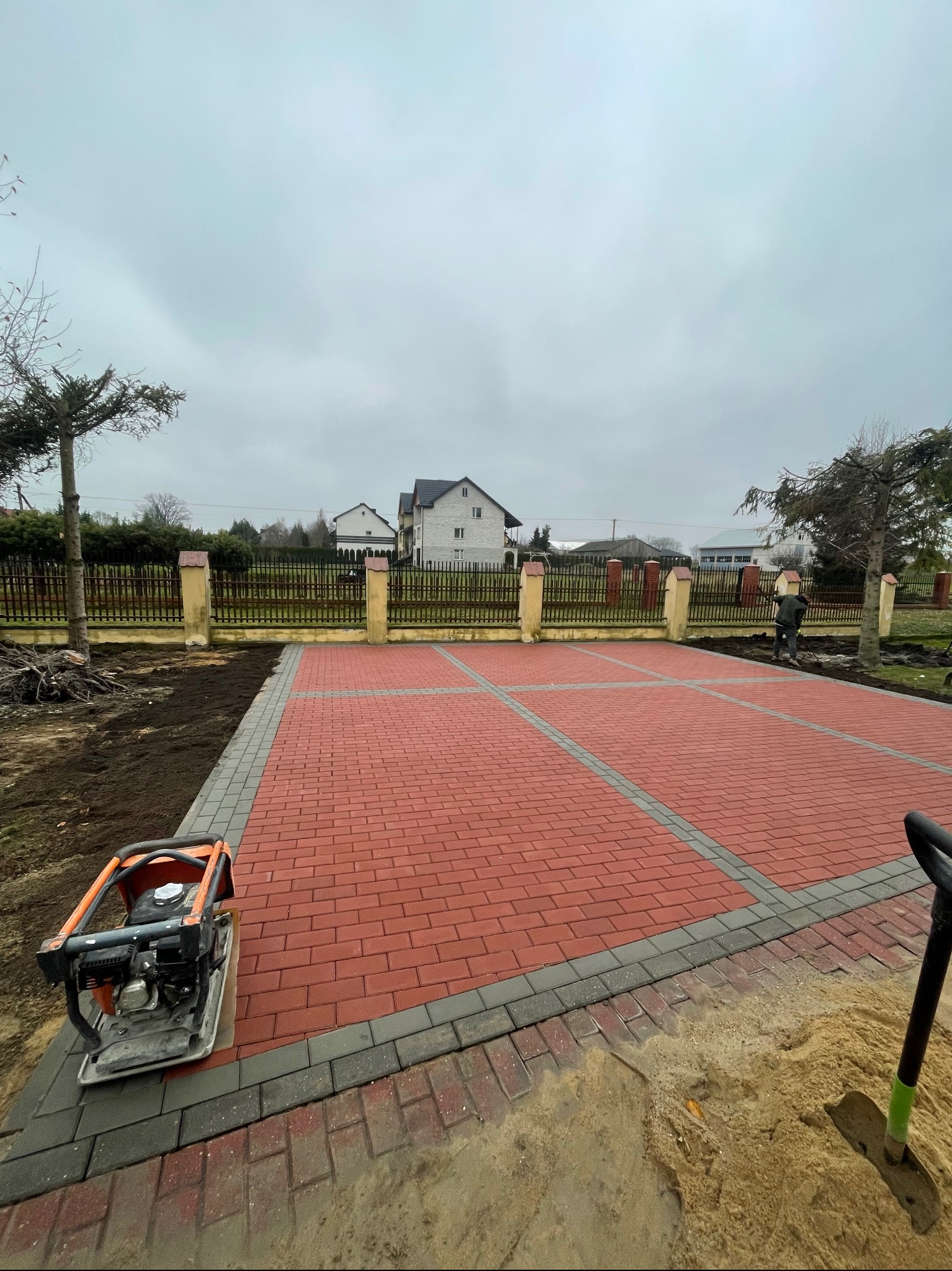 Czerwona kostka brukowa ułożona w prostokątnym wzorze, obrzeża z szarej kostki. Ubijarka i łopata w pobliżu. Tło: ogrodzenie i domy. Pochmurne niebo.
