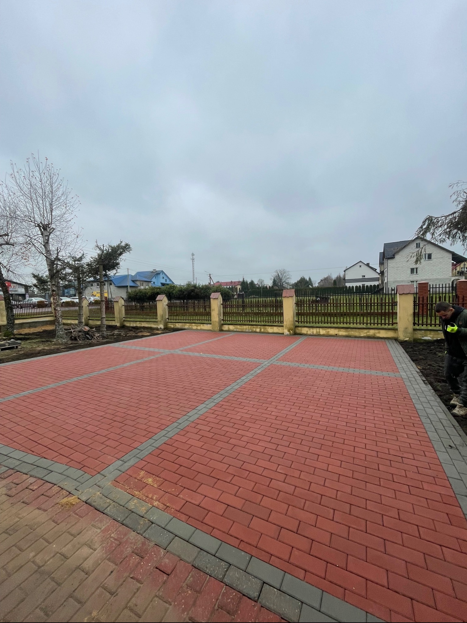 Czerwona kostka brukowa ułożona w kwadraty, oddzielone szarymi pasami. W tle ogrodzenie z drewnianymi sztachetami i budynki mieszkalne. Prace wykończeniowe w toku.