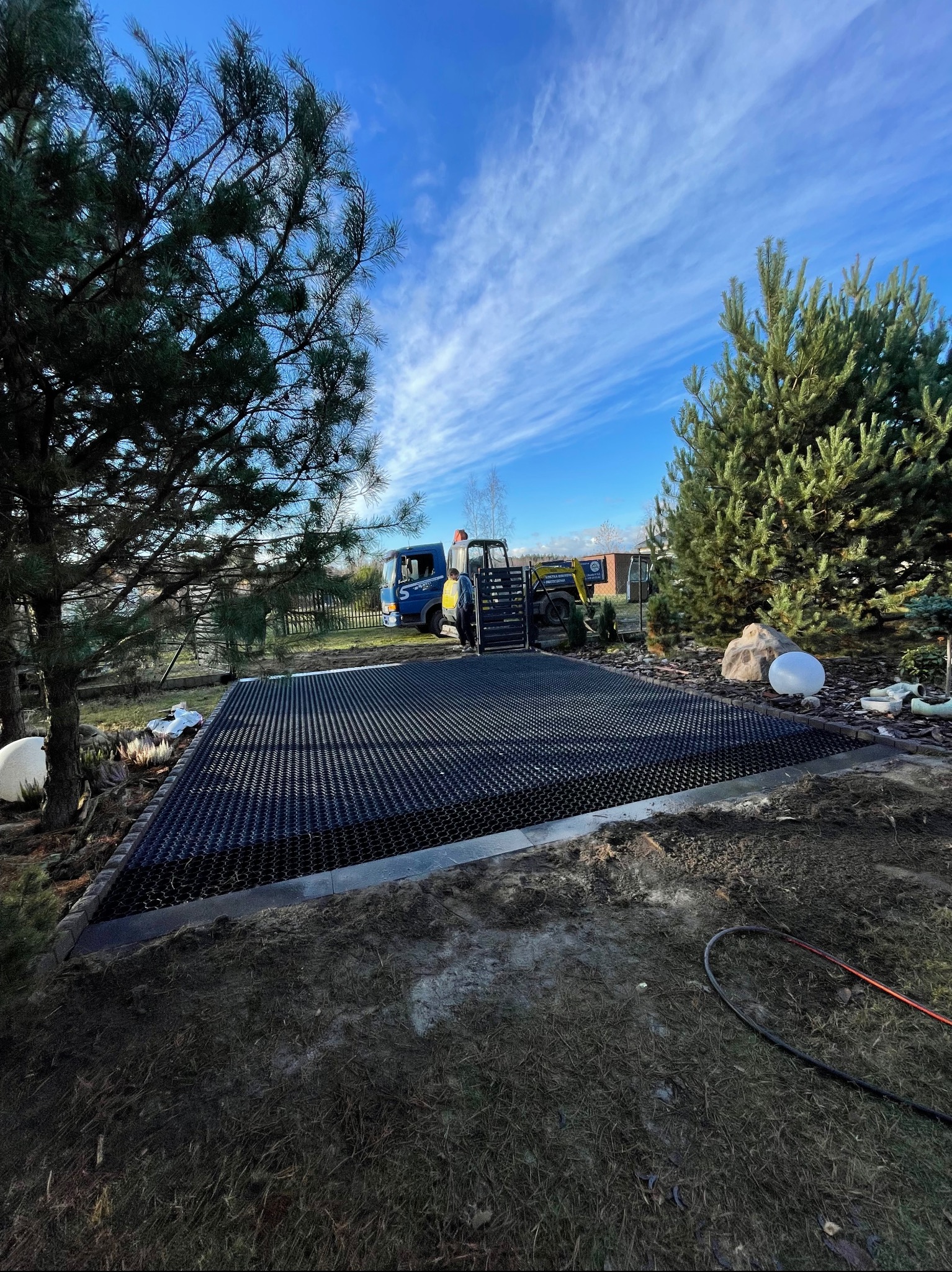 Czarna, ażurowa mata na przygotowanym podłożu, obrzeża z kostki, w tle maszyny budowlane i błękitne niebo z chmurami. Przygotowanie podłoża pod nawierzchnię.