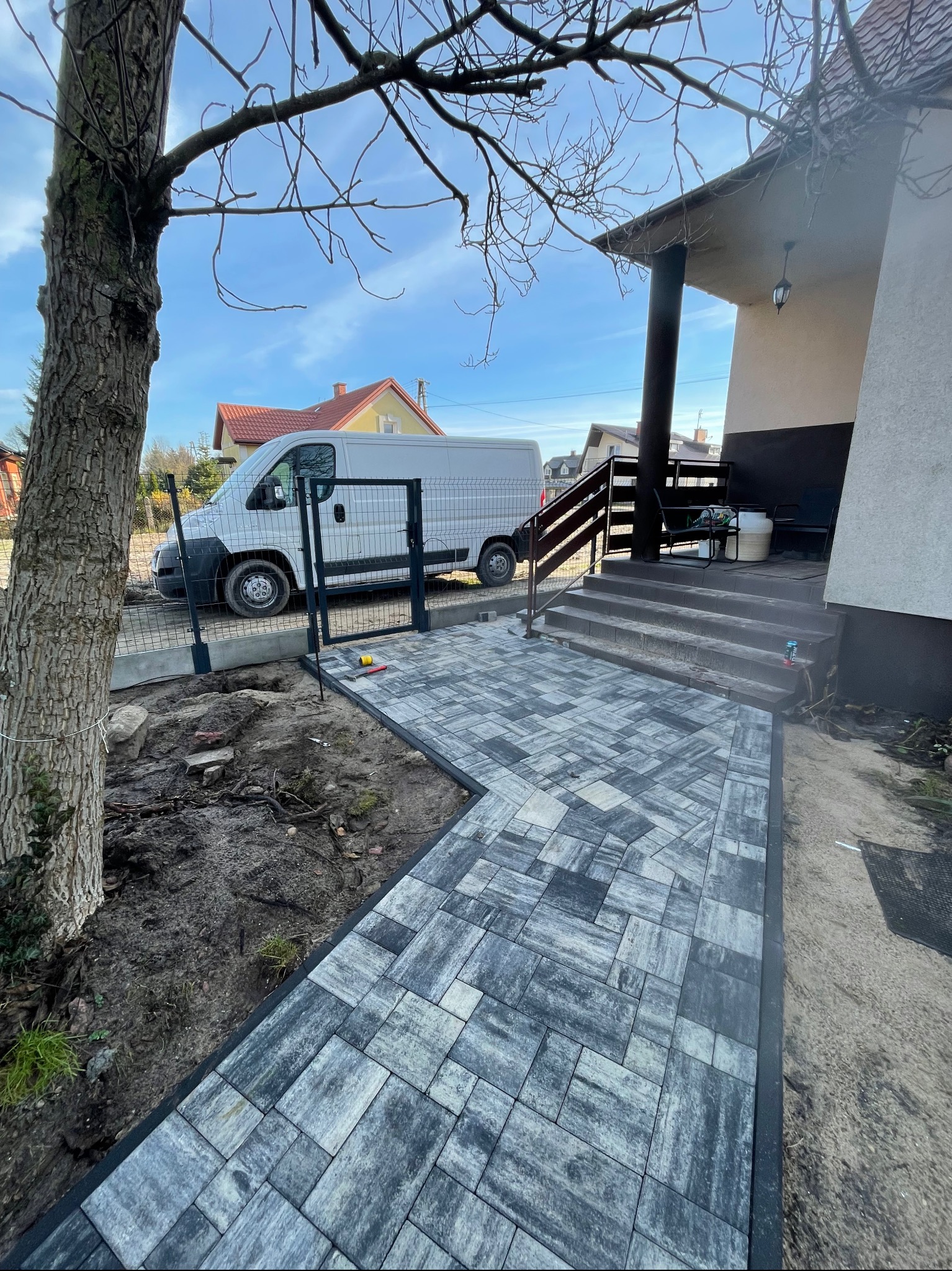 Ułożona ścieżka z kostki brukowej prowadząca do schodów wejściowych domu. W tle biały bus i ogrodzenie. Częściowo widoczne drzewo po lewej stronie.