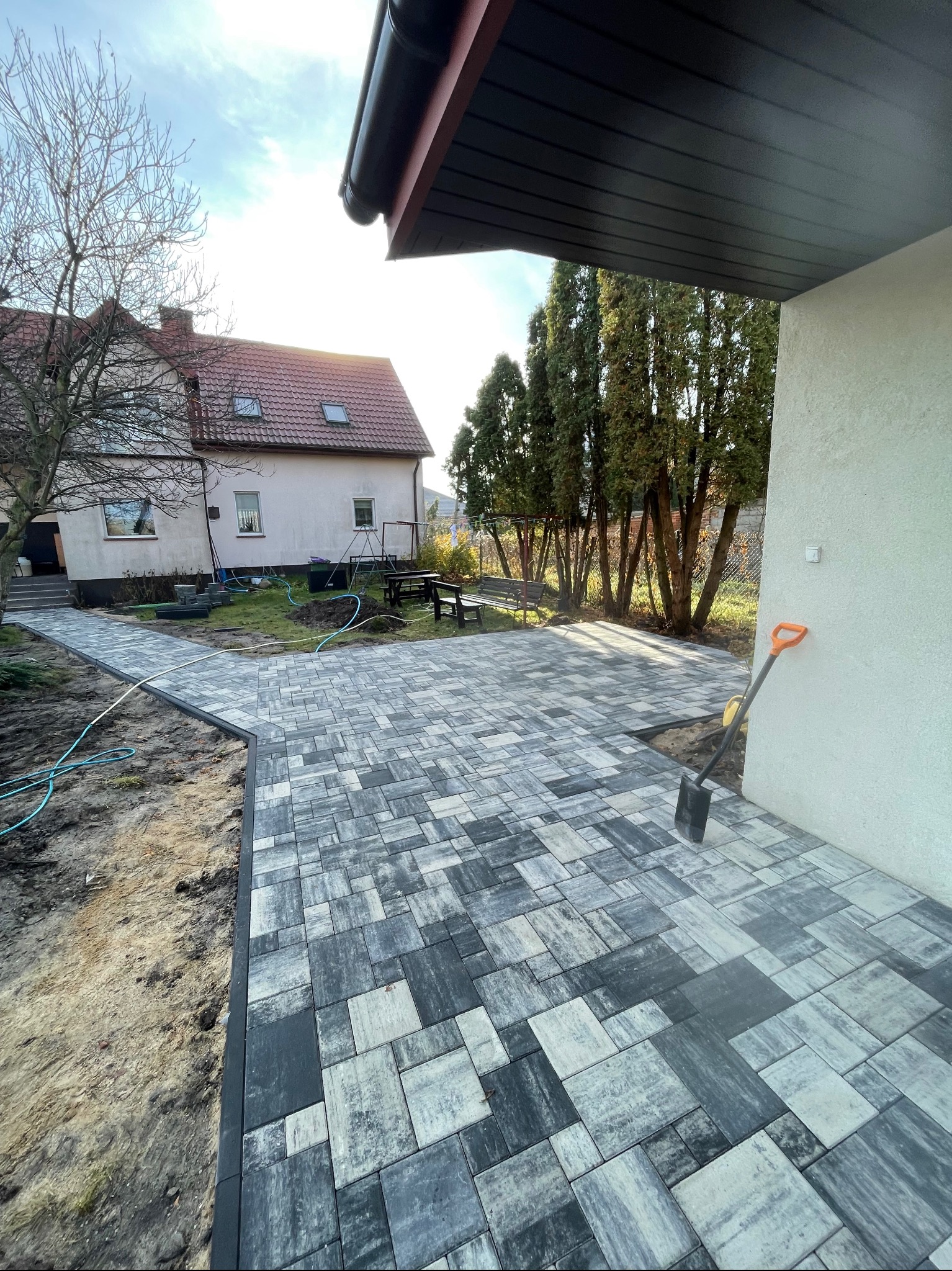 Ułożona kostka brukowa w odcieniach szarości tworzy ścieżkę i plac. W tle dom z czerwonym dachem i drzewa. Pomarańczowa łopata oparta o ścianę. Słoneczny dzień.