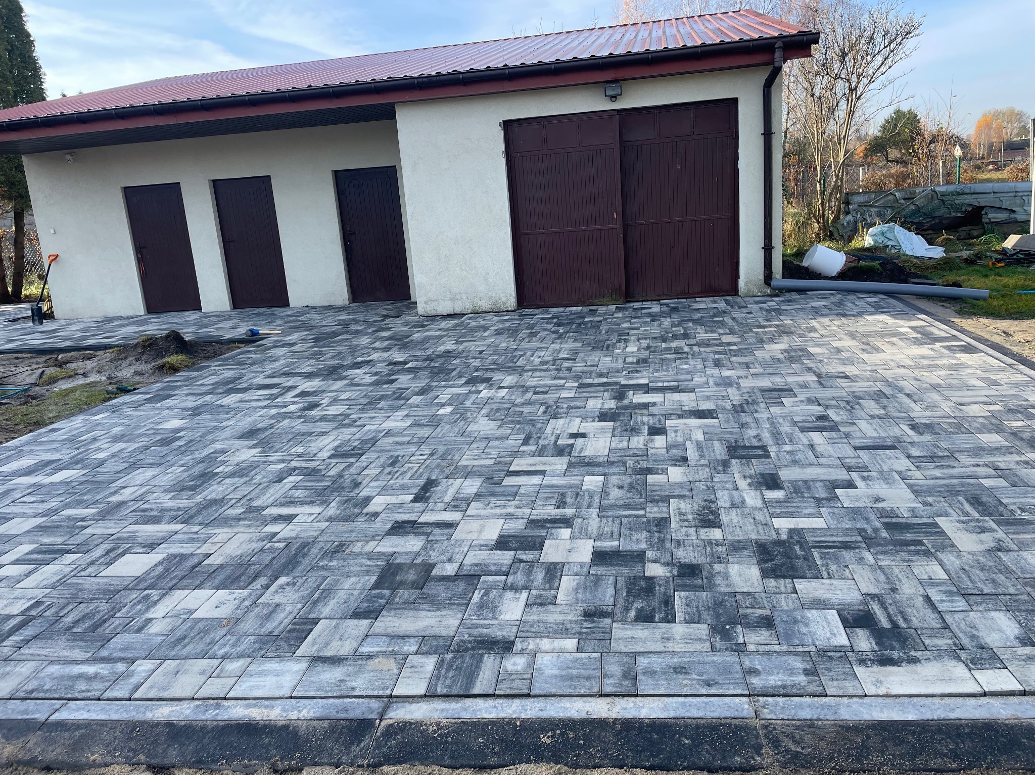 Ułożona kostka brukowa w odcieniach szarości przed budynkiem z trzema parami drzwi i bramą garażową. Widoczne obrzeża i przygotowany teren pod dalsze prace.