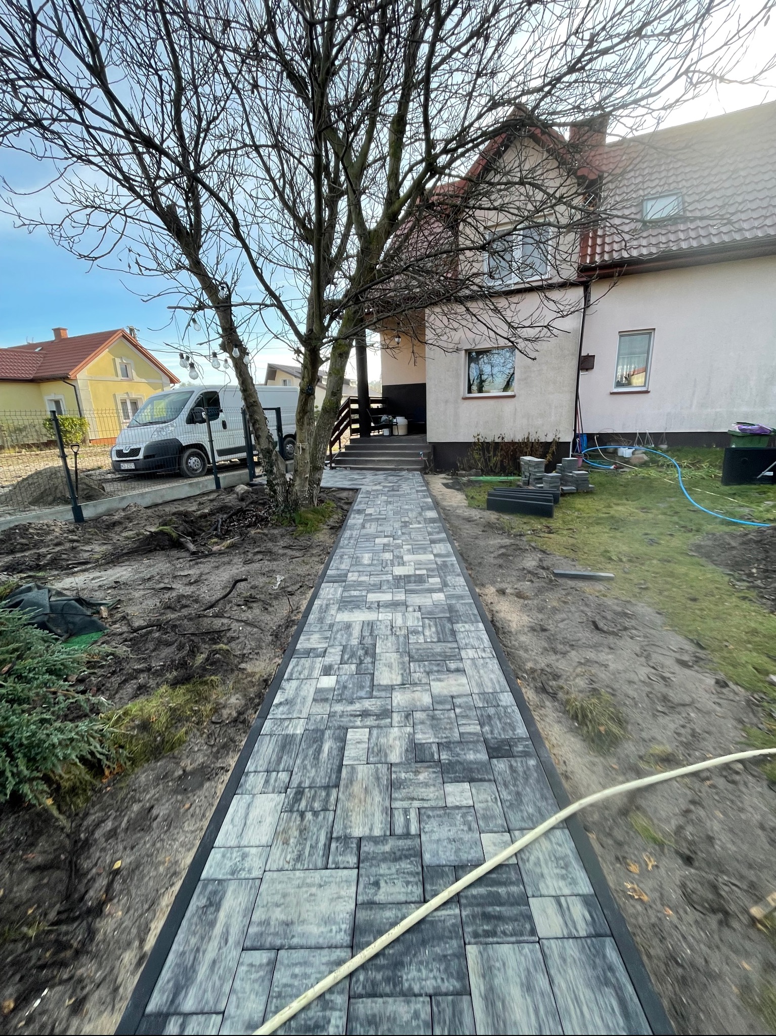 Ułożona ścieżka z szarej kostki brukowej prowadząca do domu, z widocznym samochodem dostawczym w tle i elementami ogrodu w trakcie aranżacji.