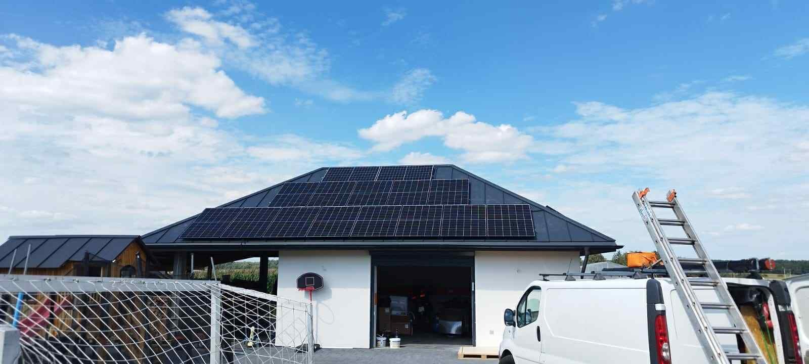 Dom z panelami fotowoltaicznymi na dachu, drabina oparta o samochód dostawczy. Widoczne błękitne niebo z chmurami i fragment boiska do koszykówki.