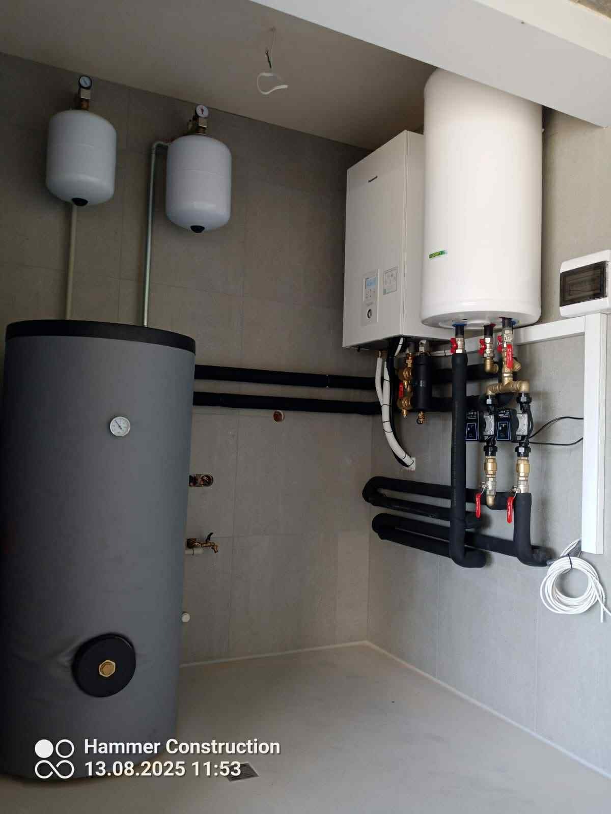 Kompletna instalacja grzewcza z zasobnikiem, kotłem i rurami w nowym pomieszczeniu. Widoczne czarne, izolowane rury i białe zbiorniki wyrównawcze.