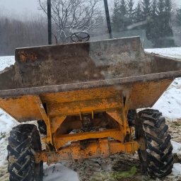 Firma usługowa Marcin Krasowski - Żółta wywrotka budowlana z dużymi oponami, widziana z tyłu, stoi na ośnieżonym terenie. W tle drzewa i mgła. Widok zza szyby.