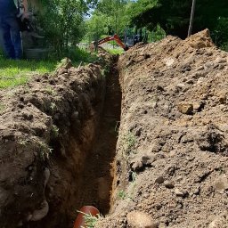 Firma usługowa Marcin Krasowski - Wykopany rów w ziemi z pomarańczową rurą na dnie, w tle minikoparka i pracownik w zielonej koszulce. Prace ziemne w słoneczny dzień.