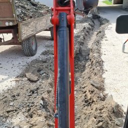 Firma usługowa Marcin Krasowski - Czerwona koparka w trakcie wykonywania wykopu pod instalacje na żwirowym terenie, widok z kabiny operatora, w tle budynek gospodarczy i przyczepa z ziemią.