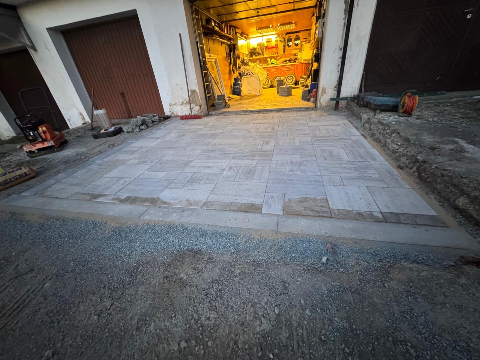 Ułożona kostka brukowa na podjeździe przed garażem, widoczny zagęszczarka i przygotowane podłoże. Etap prac wykończeniowych.