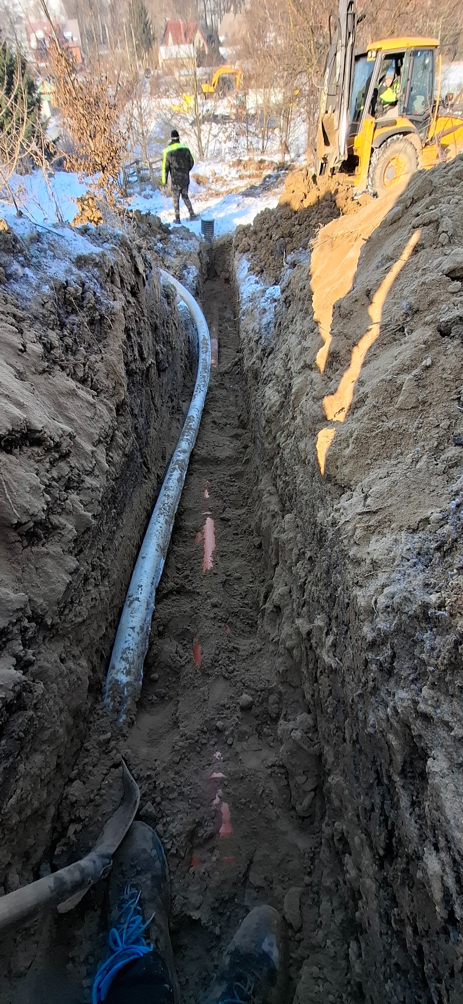 Wykop z ułożoną rurą, widoczne oznaczenia geodezyjne, ziemia, w tle koparka i pracownik. Prace ziemne w terenie zabudowanym, zimowa sceneria.