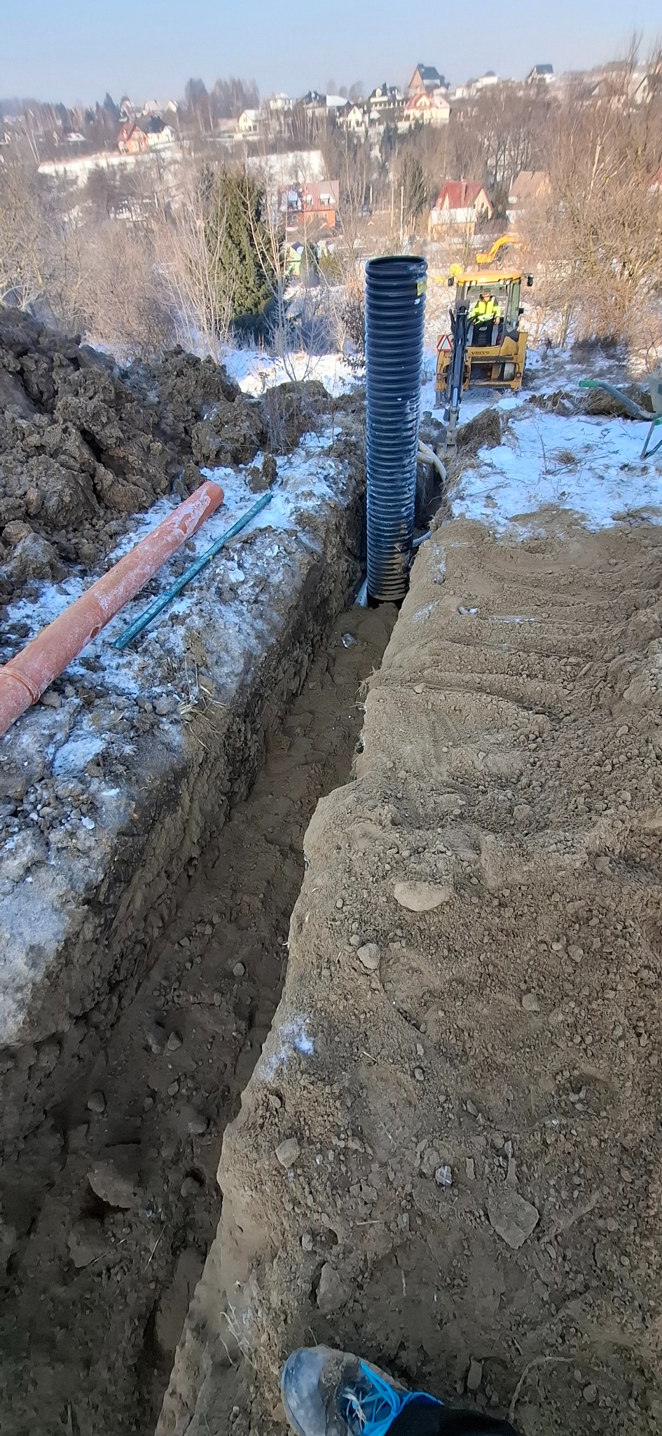 Wykop pod rurę drenażową z widocznym traktorem w tle. Rura karbowana, pionowa. Ziemia częściowo pokryta śniegiem. Widok na zabudowania w oddali.