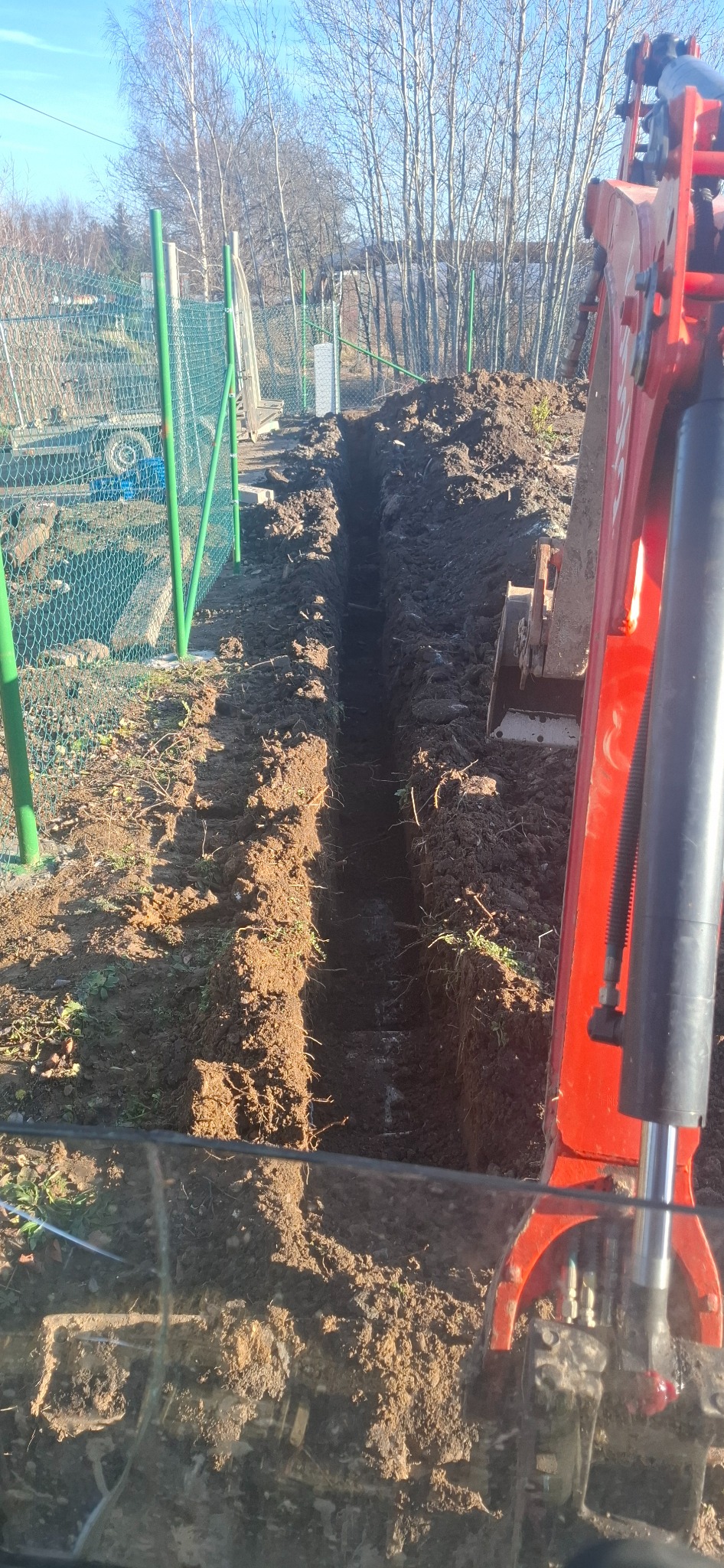 Wąski wykop w ziemi, wykonany prawdopodobnie pod instalacje, widoczny fragment koparki i ogrodzenie z siatki w tle, słoneczny dzień.