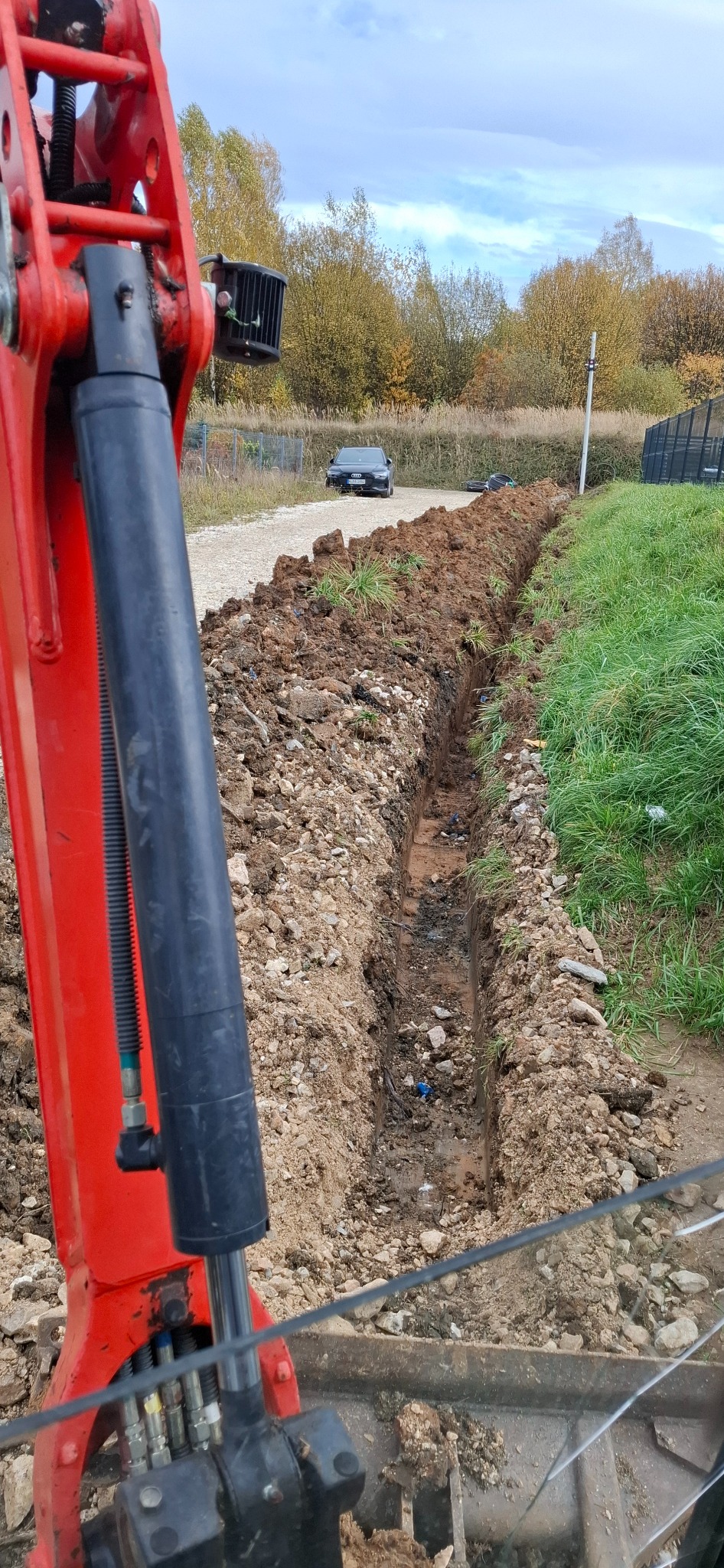 Wykopany wąski rów w ziemi, widoczny z perspektywy operatora czerwonej minikoparki. W tle droga gruntowa i dwa samochody osobowe.