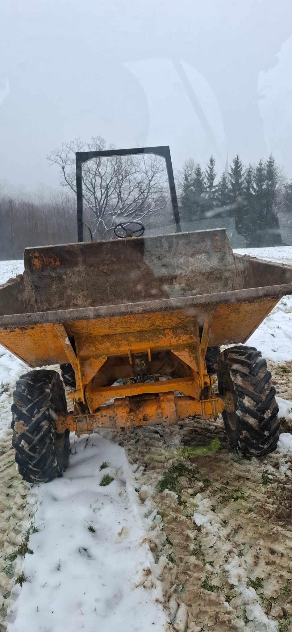 Żółta wywrotka budowlana z dużymi oponami, widziana z tyłu, stoi na ośnieżonym terenie. W tle drzewa i mgła. Widok zza szyby.