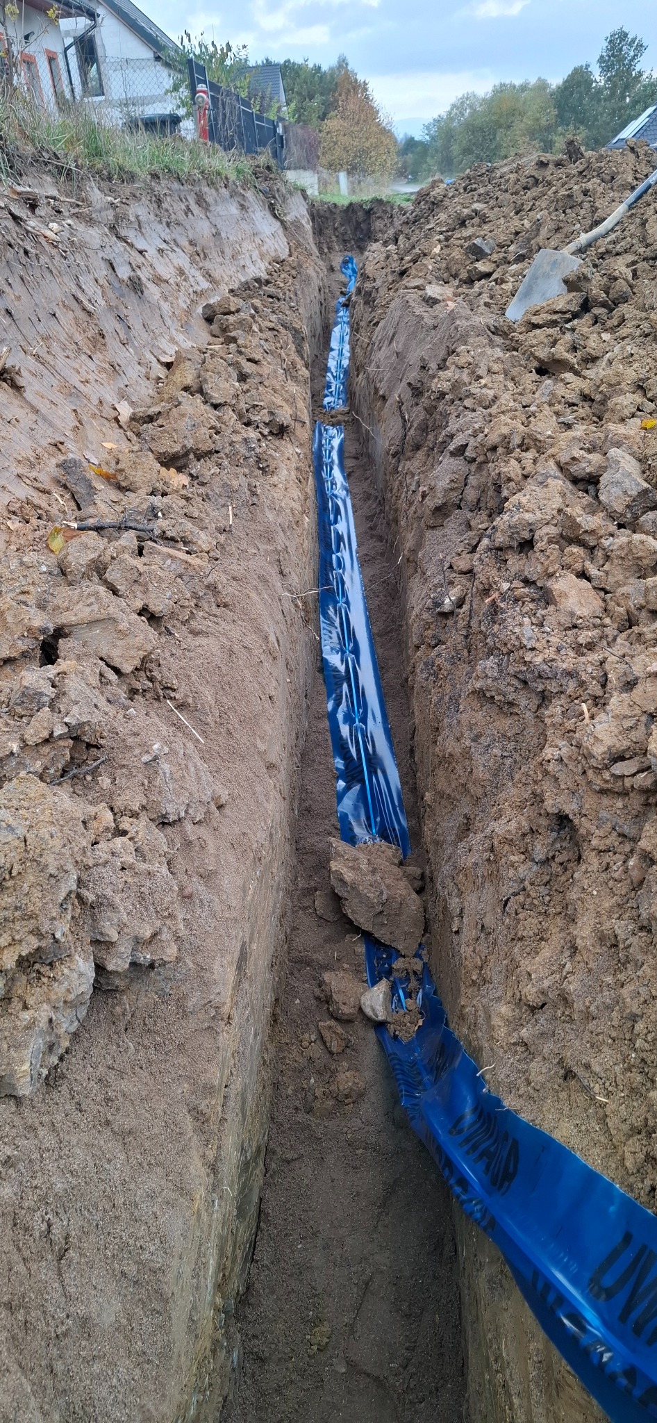 Wąski wykop w ziemi z niebieską folią ochronną na dnie, prawdopodobnie pod instalację. Widoczna ziemia, narzędzia i fragment zabudowań w tle.