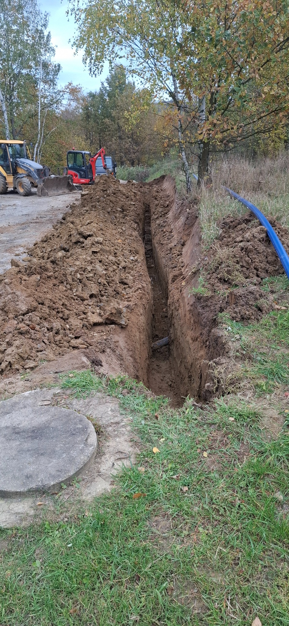 Wykopany wąski rów w ziemi, z widoczną rurą na dnie. W tle maszyny budowlane: koparka i ładowarka. Na pierwszym planie betonowy element. Jesienne otoczenie.