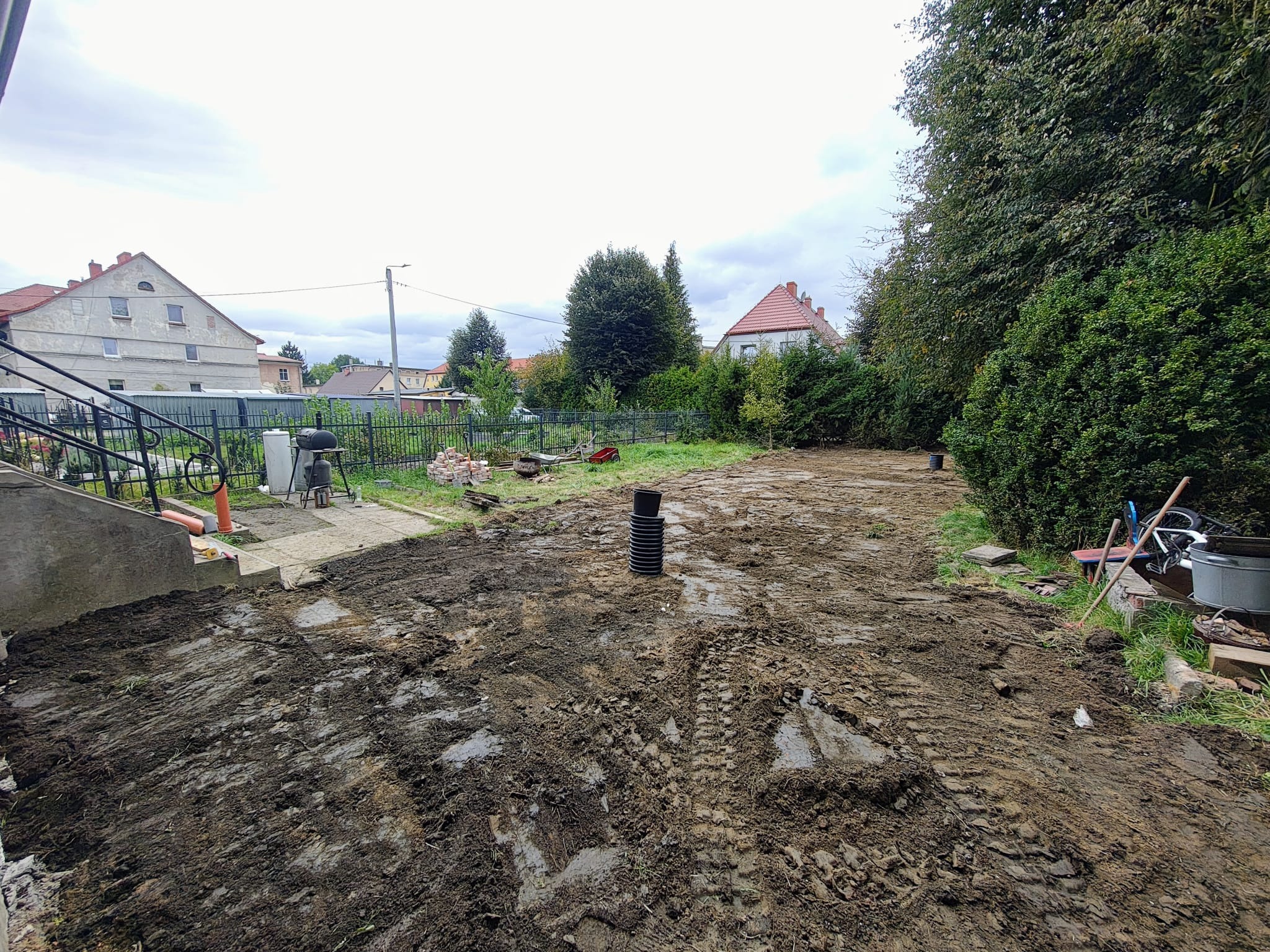 Przygotowany teren pod budowę w Krosnowicach. Widoczne ślady opon i czarna rura drenażowa. W tle budynki mieszkalne i zieleń.