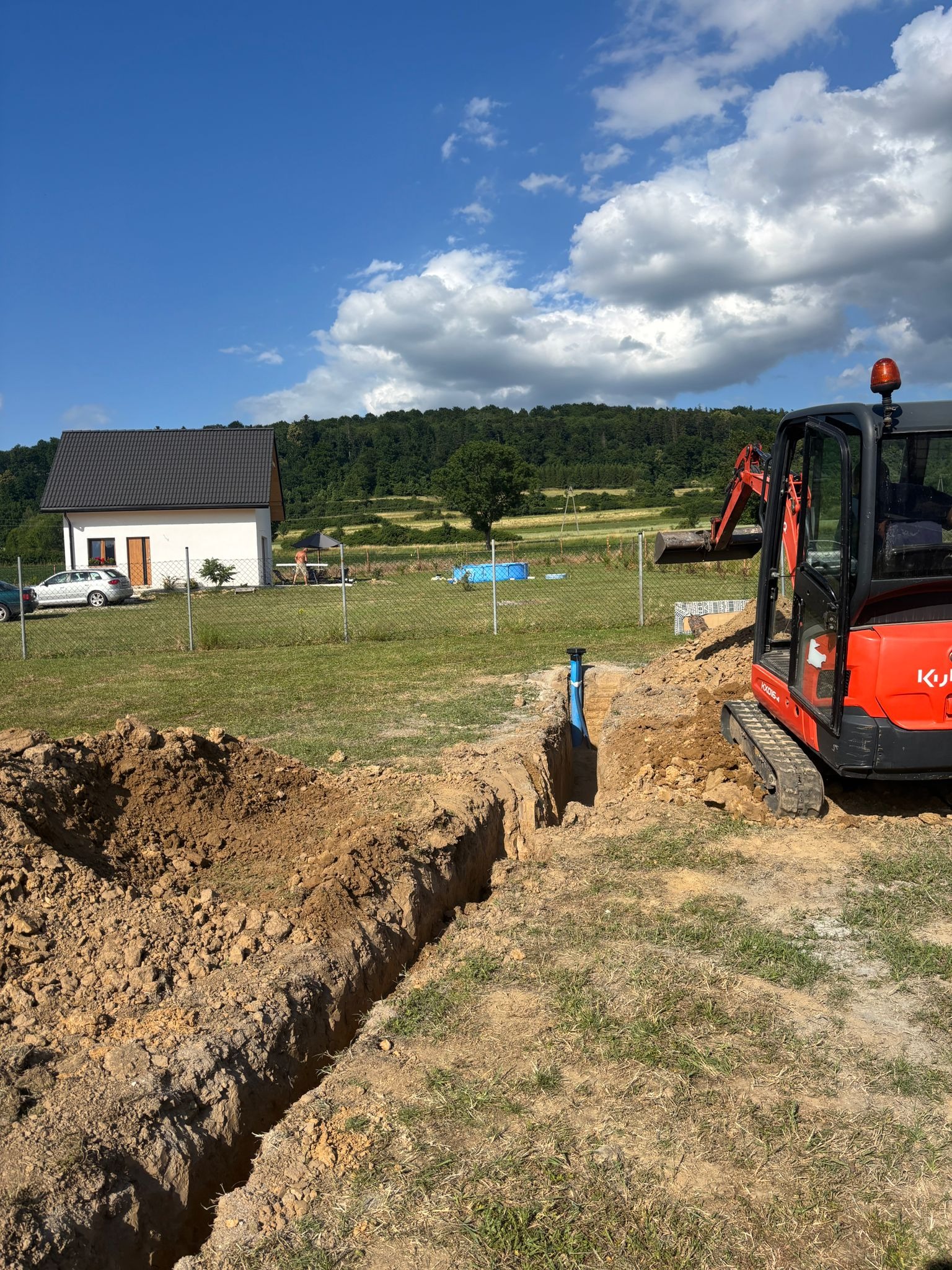 Wykopany wąski rów z wystającą niebieską rurą, obok stoi pomarańczowa mini koparka Kubota. W tle dom jednorodzinny i zalesione wzgórze pod błękitnym niebem.