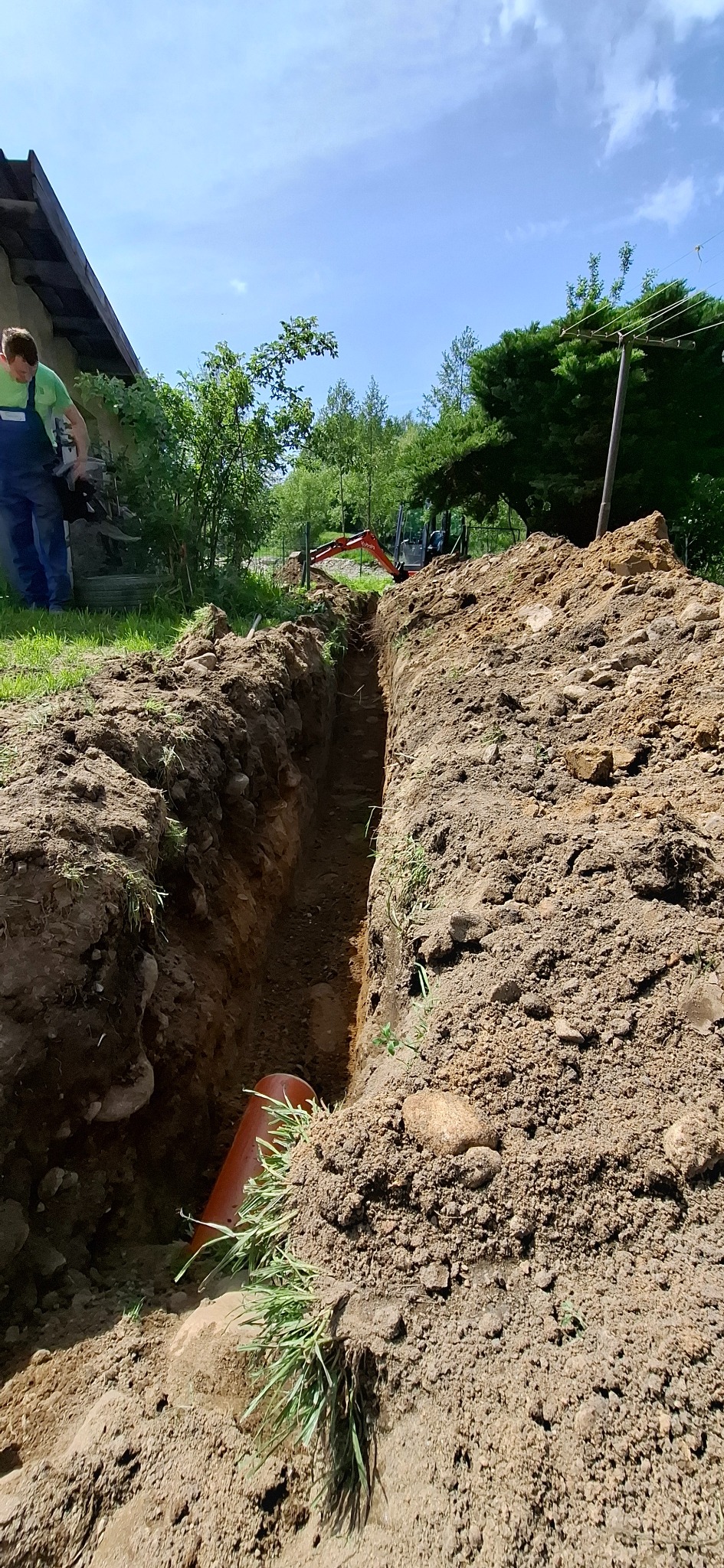 Wykopany rów w ziemi z pomarańczową rurą na dnie, w tle minikoparka i pracownik w zielonej koszulce. Prace ziemne w słoneczny dzień.