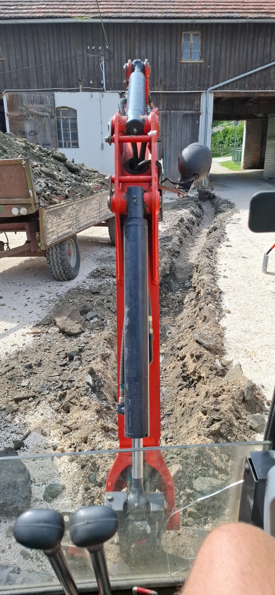 Czerwona koparka w trakcie wykonywania wykopu pod instalacje na żwirowym terenie, widok z kabiny operatora, w tle budynek gospodarczy i przyczepa z ziemią.