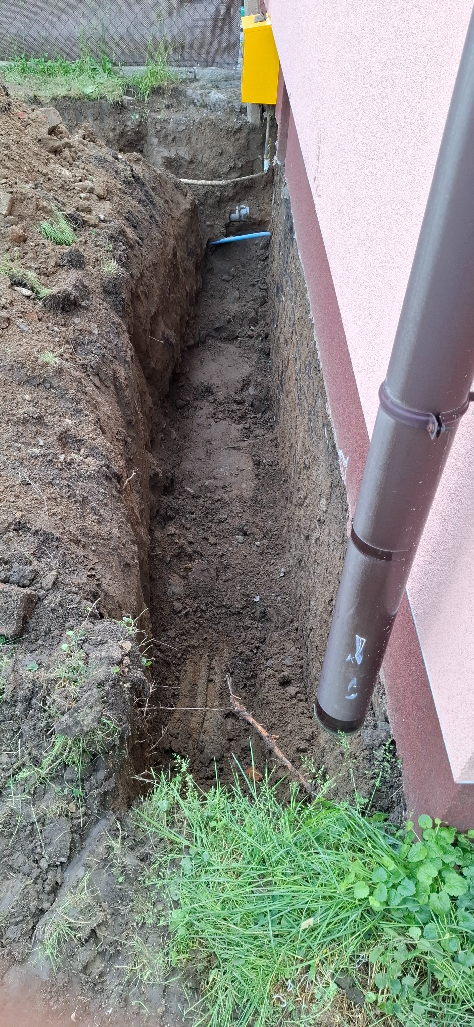 Wąski wykop w ziemi wzdłuż ściany budynku z rurą. Widoczne ślady po szpadlu. Ziemia mokra, gliniasta. Na pierwszym planie brązowa rynna i zielona trawa.