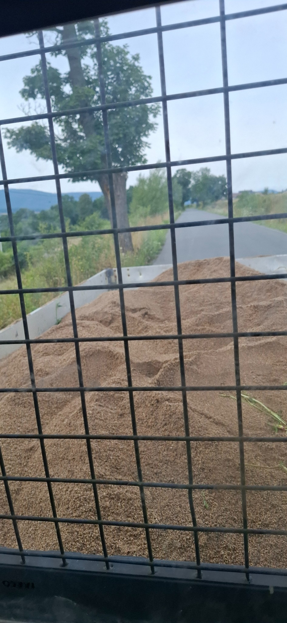 Piasek w skrzyni pojazdu, widok przez kratę ochronną. W tle droga i drzewa. Może dotyczyć transportu materiałów budowlanych lub niwelacji terenu.