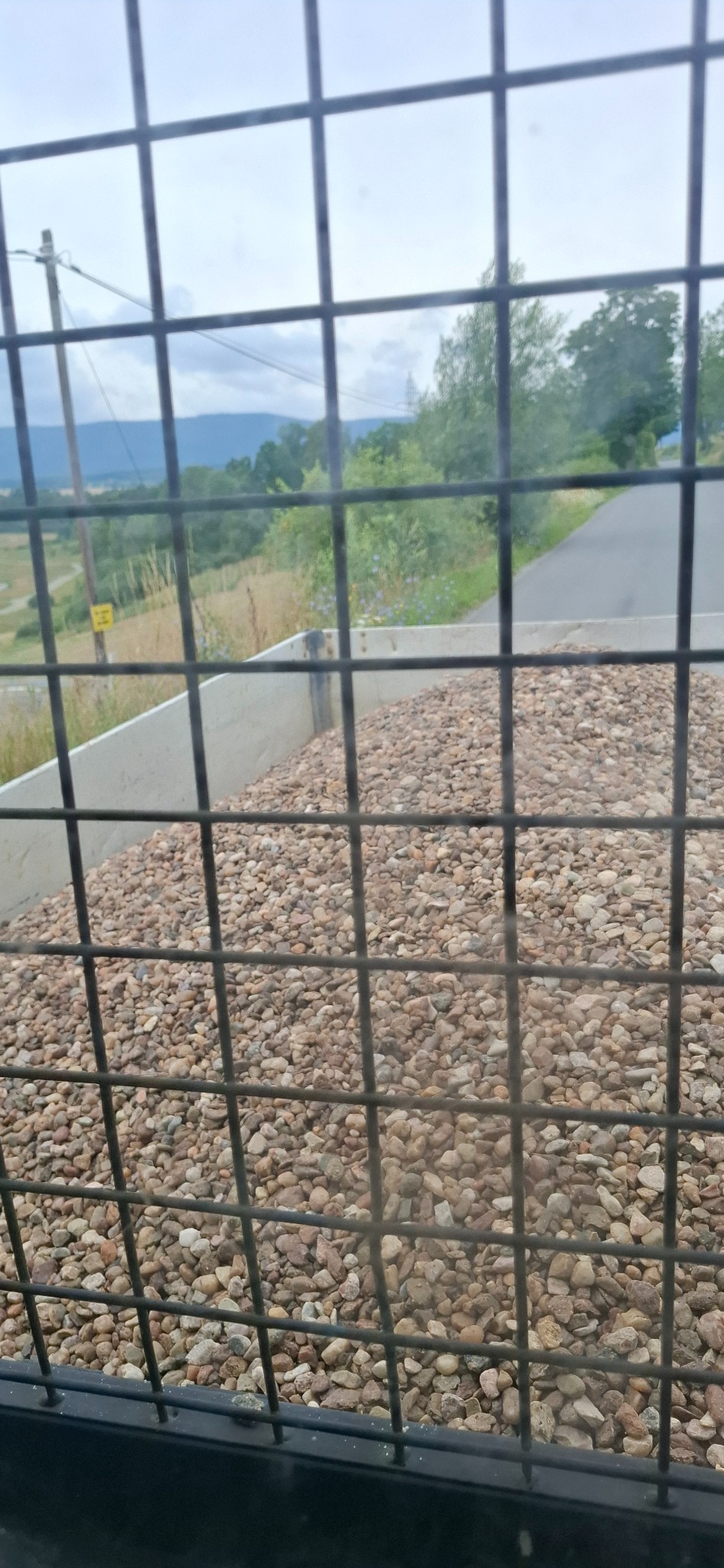 Kopalnia żwiru widziana przez kratę. W tle droga i krajobraz. Żwir w betonowej skrzyni. Widok z kabiny maszyny budowlanej.