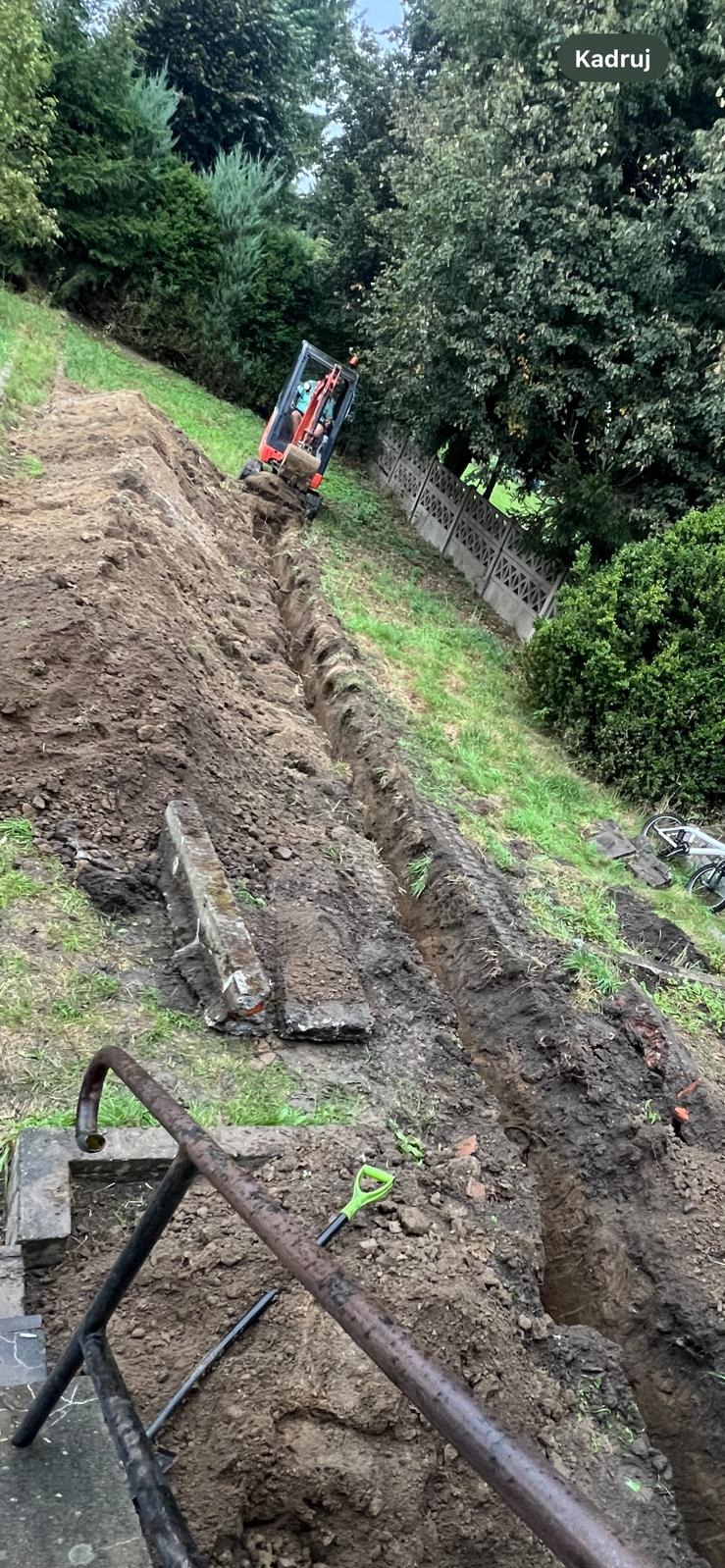 Wykopany rów na zboczu, z widocznym minikoparką na górze i fragmentem metalowej konstrukcji w dole kadru. Ślady opon wzdłuż wykopu.