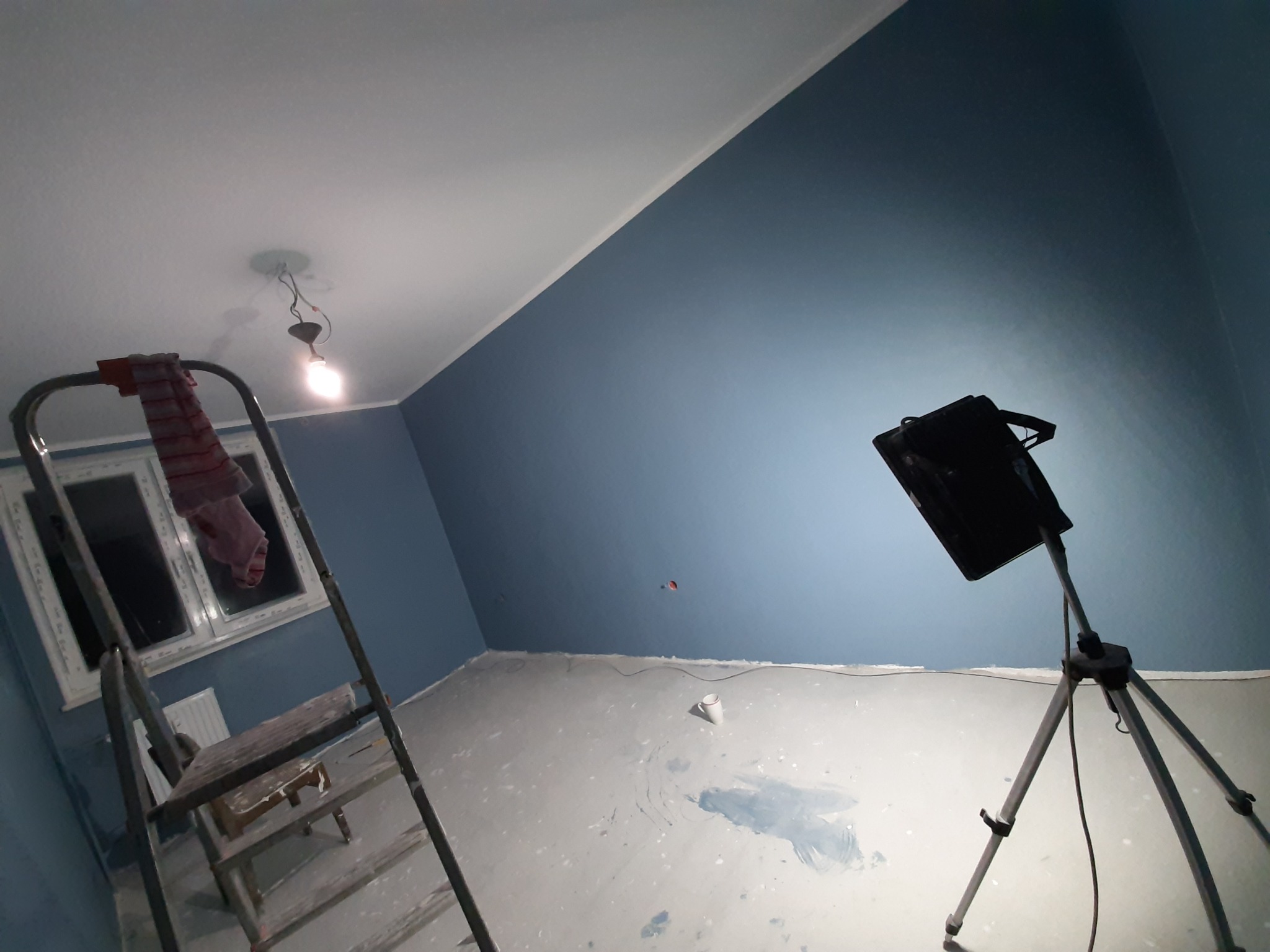 Świeżo pomalowane na niebiesko ściany w pokoju z drabiną, lampą na statywie i widocznymi śladami farby na podłodze. Widok na okno.