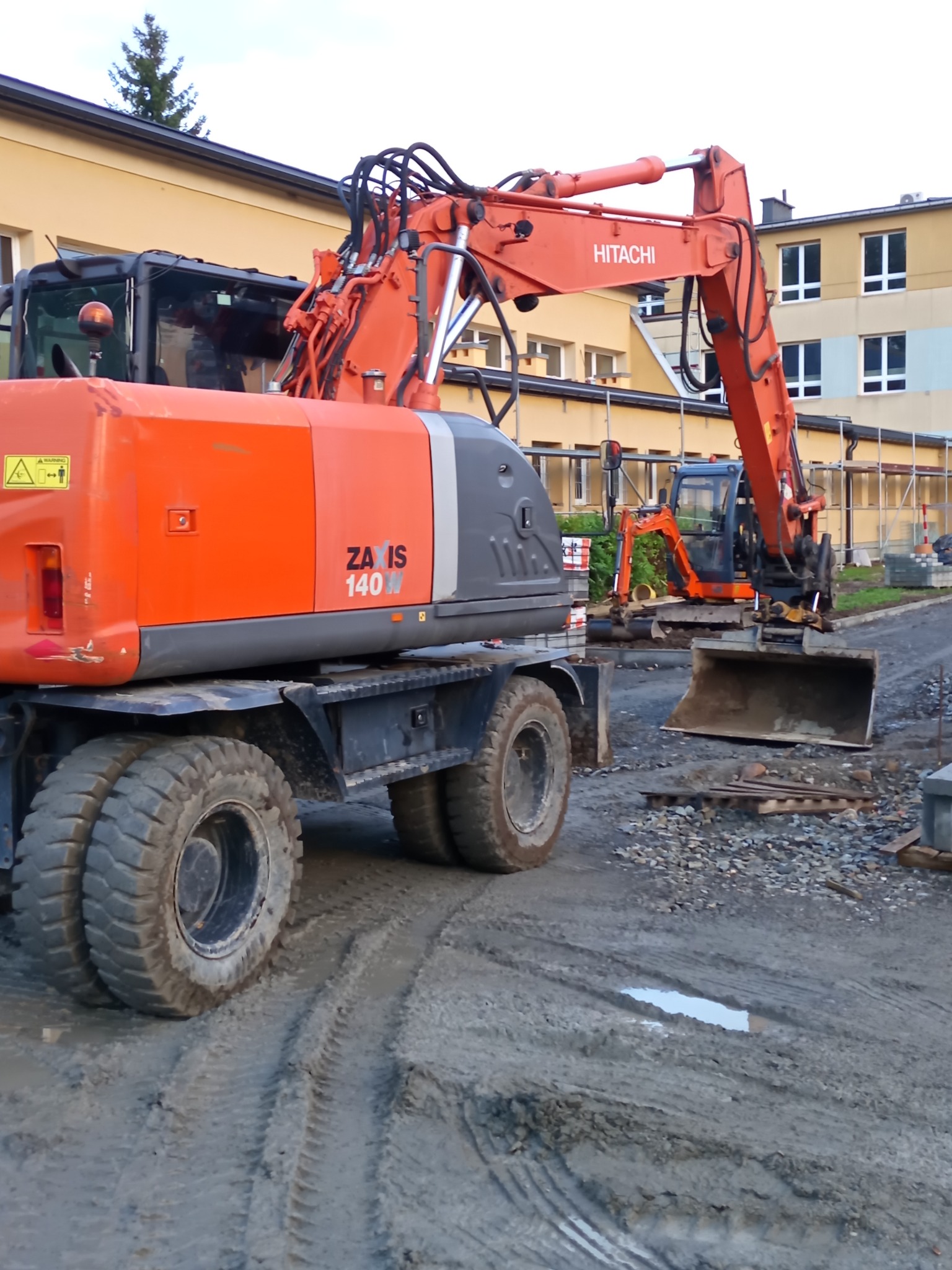 Pomarańczowa koparka Hitachi Zaxis 140W na błotnistym terenie budowy, w tle budynek i druga, mniejsza koparka. Widoczne ślady opon i kałuża.