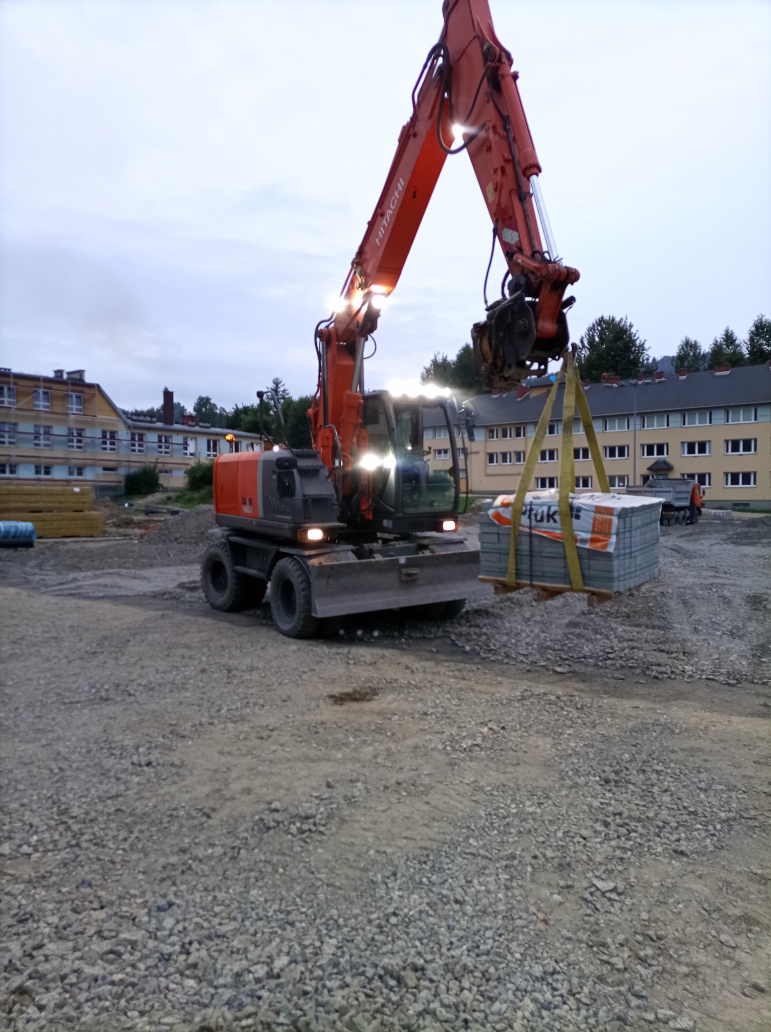 Pomarańczowa koparka kołowa Hitachi podnosi paletę kostki brukowej na placu budowy. W tle budynki i ciężarówka. Oświetlenie robocze włączone.