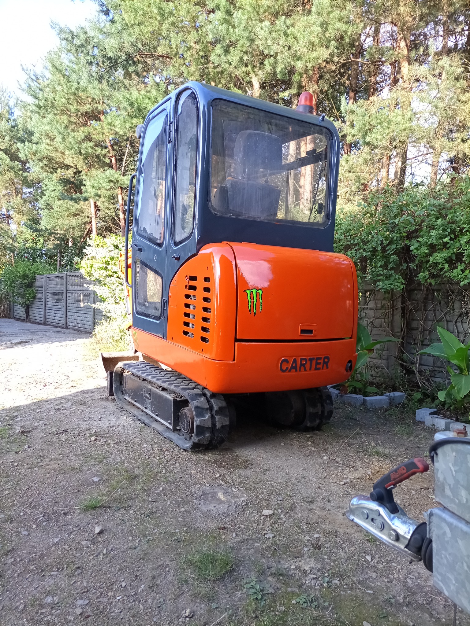 Pomarańczowa mini koparka Carter z zielonym logo Monster Energy, stoi na żwirowym podłożu, w tle drzewa i ogrodzenie. Widoczny zaczep przyczepy.