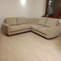 BRATEX Przemysław Bratkowski - Beżowy, narożny, tapicerowany kanapa z subtelnymi złotymi nóżkami, ustawiona na jasnej podłodze. Widoczne przeszycia i detale wykończenia. Nowoczesny design.