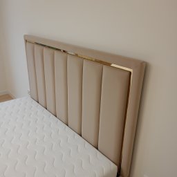 BRATEX Przemysław Bratkowski - Beżowe, tapicerowane wezgłowie łóżka z dekoracyjną, złotą ramką, widoczne z boku, z materacem. Minimalistyczny design i neutralna kolorystyka.