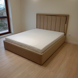 BRATEX Przemysław Bratkowski - Beżowe, tapicerowane łóżko z wysokim wezgłowiem, wykończonym złotymi akcentami, stoi na drewnianej podłodze w jasnym pokoju. Widok z perspektywy.