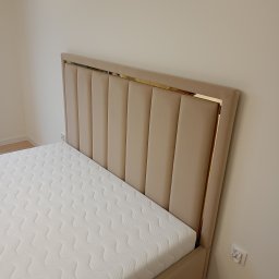 BRATEX Przemysław Bratkowski - Beżowe łóżko tapicerowane z wysokim, pionowo pikowanym wezgłowiem, wykończonym złotą ramką. Biały materac z delikatnym wzorem. Minimalistyczne wnętrze.