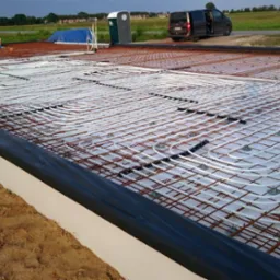 Przygotowanie do wylewki z ogrzewaniem podłogowym: białe rurki grzewcze ułożone na siatce zbrojeniowej, widoczny szalunek i grunt w tle.