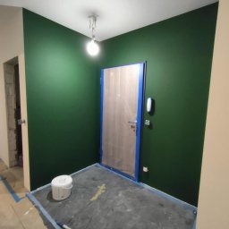 YULIIA SIDORENKO - Ściana pomalowana na zielono, zabezpieczone folią drzwi i podłoga. Widoczny biały sufit i lampa. Kątowe ujęcie. Częściowo widoczne wiadro z farbą i wałek.