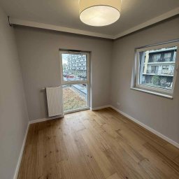 YULIIA SIDORENKO - Jasne, drewniane panele podłogowe w pokoju z szarymi ścianami, białymi listwami przypodłogowymi i dwoma oknami z widokiem na zieleń i budynki. Okrągła lampa sufitowa.