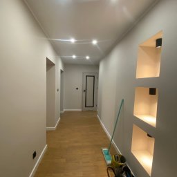YULIIA SIDORENKO - Wykończony korytarz z drewnianą podłogą, wnękami z oświetleniem i świeżo pomalowanymi ścianami w odcieniach szarości. Widoczne akcesoria do sprzątania.