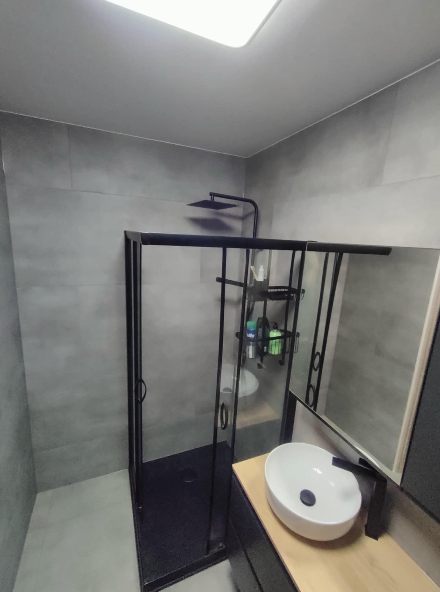 Nowoczesna łazienka z kabiną prysznicową typu walk-in z czarnymi ramami i deszczownicą. Umywalka nablatowa na drewnianym blacie. Szare płytki na ścianach i podłodze.