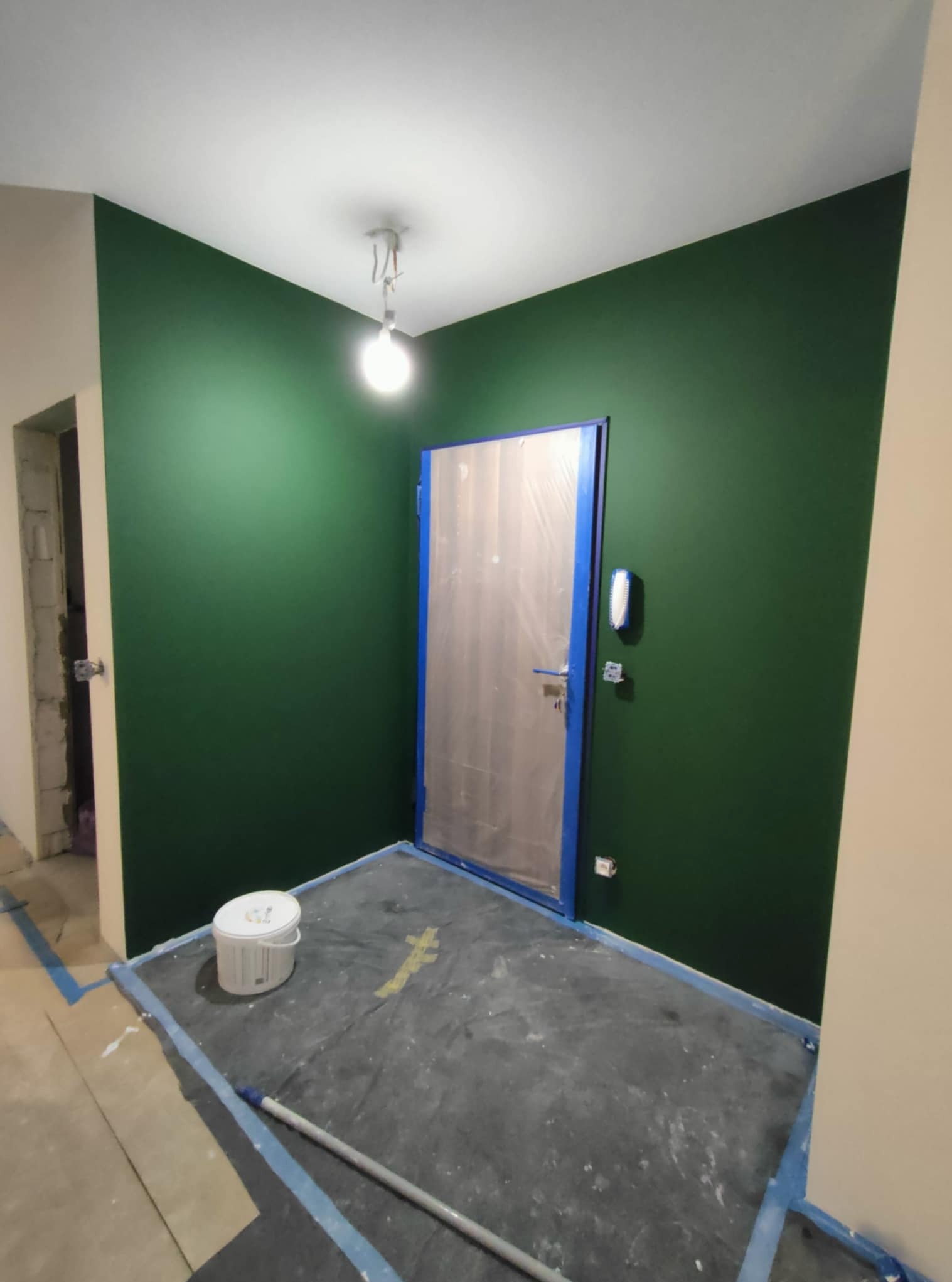 Ściana pomalowana na zielono, zabezpieczone folią drzwi i podłoga. Widoczny biały sufit i lampa. Kątowe ujęcie. Częściowo widoczne wiadro z farbą i wałek.