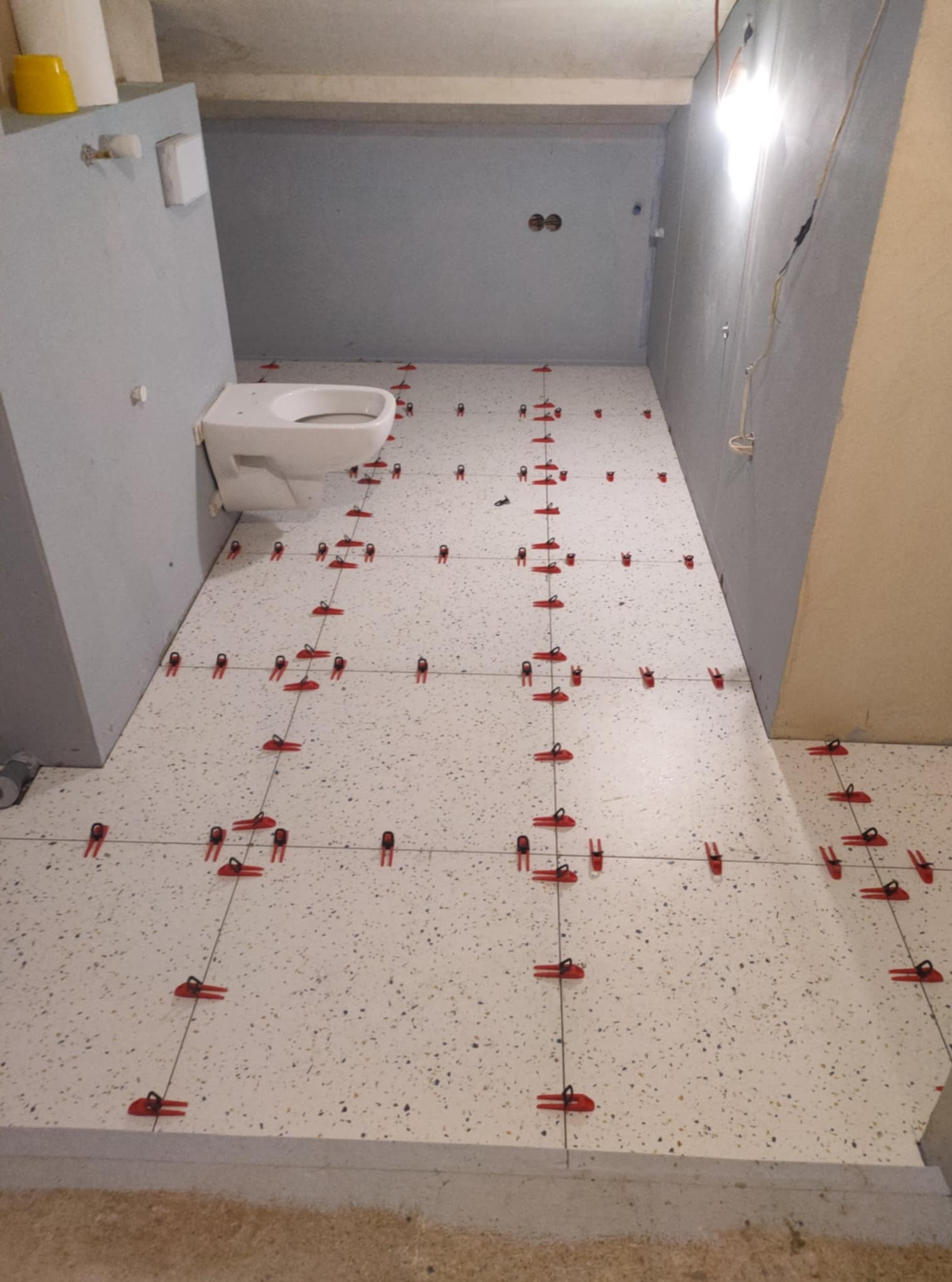 Biała podłoga w łazience z terazzo, z czerwonymi klipsami poziomującymi płytki, częściowo zamontowana toaleta, szare ściany w trakcie wykańczania.