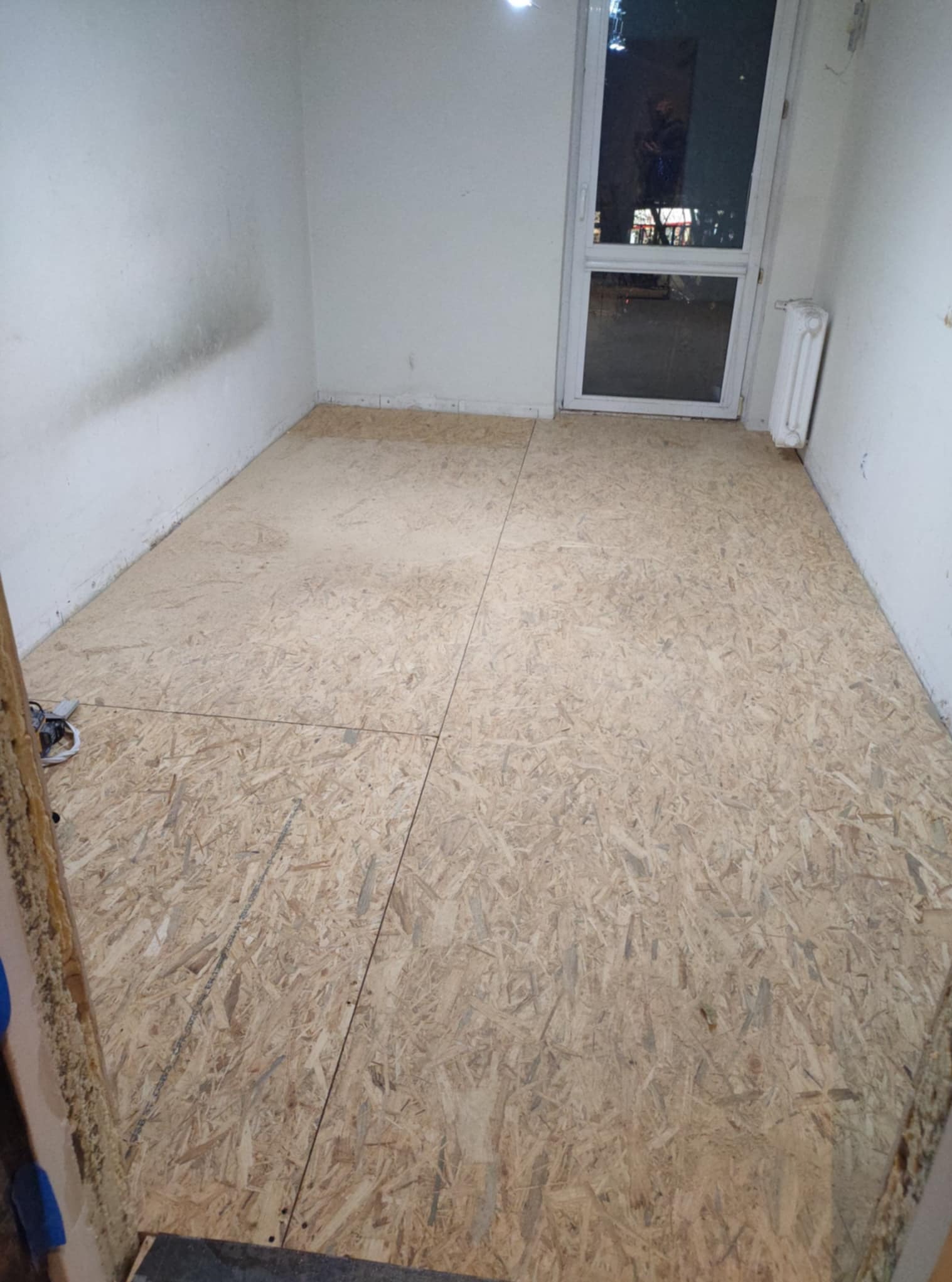 Przygotowanie podłoża: płyty OSB ułożone na podłodze w pustym pomieszczeniu z oknem i grzejnikiem. Widoczne ślady po starej podłodze na ścianach.